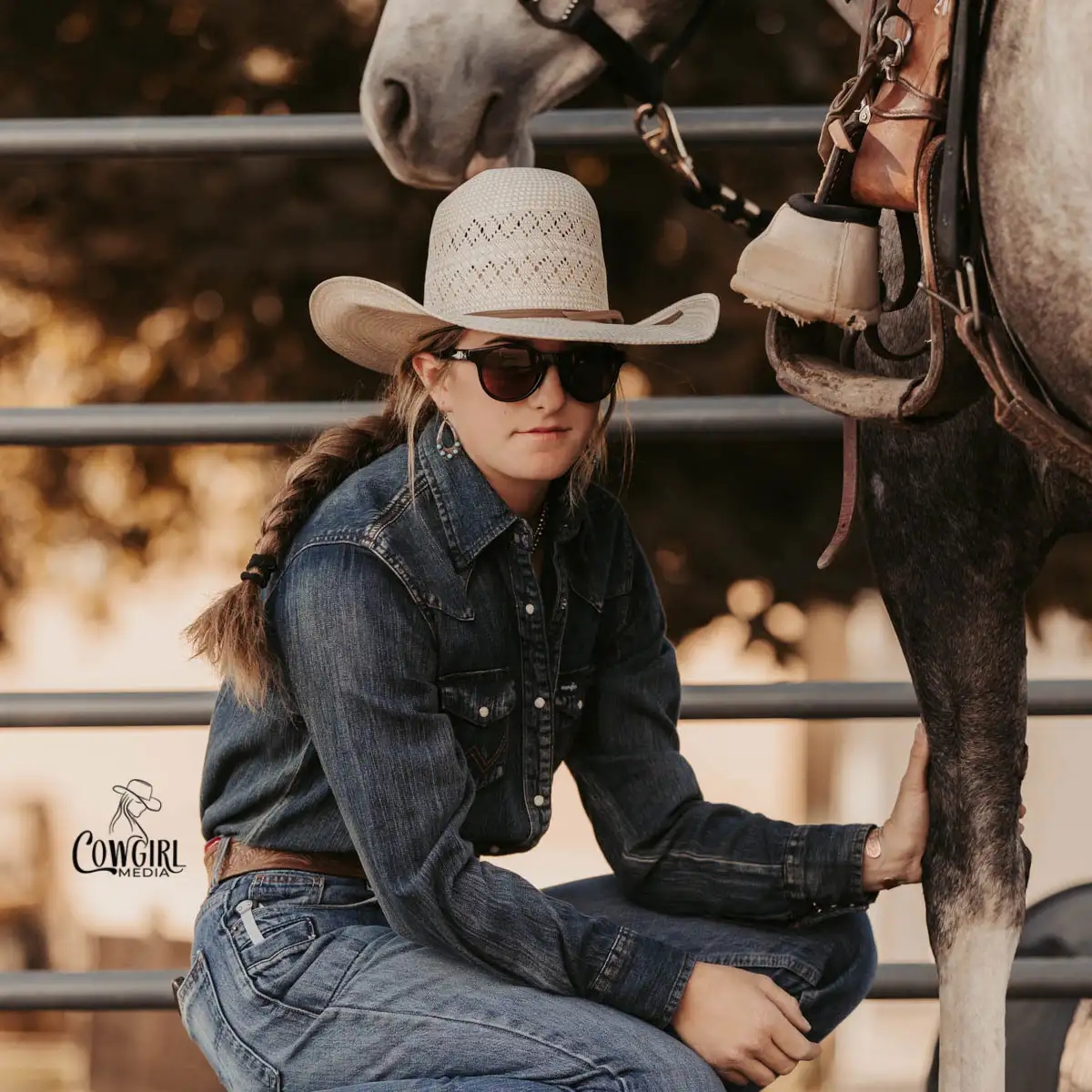 Cowgirl wearing sunglasses squatting down next to her horse with her hand on his leg